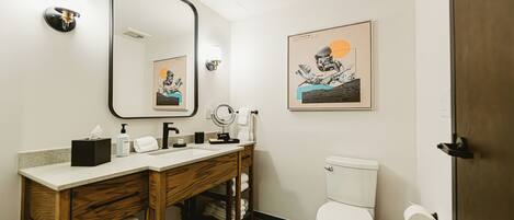 A modern bathroom with a wooden vanity, a large mirror, a toilet, and artwork on the walls.