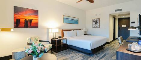 A hotel room with a bed, a desk, a chair, a table with flowers, a ceiling fan, and artwork on the walls.