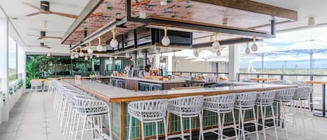 Un espacio de bar moderno con una barra alargada, taburetes blancos y ventiladores de techo.