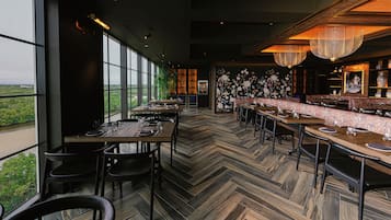 Um restaurante com chão em padrão herringbone, grandes janelas e área de bar com um painel floral.