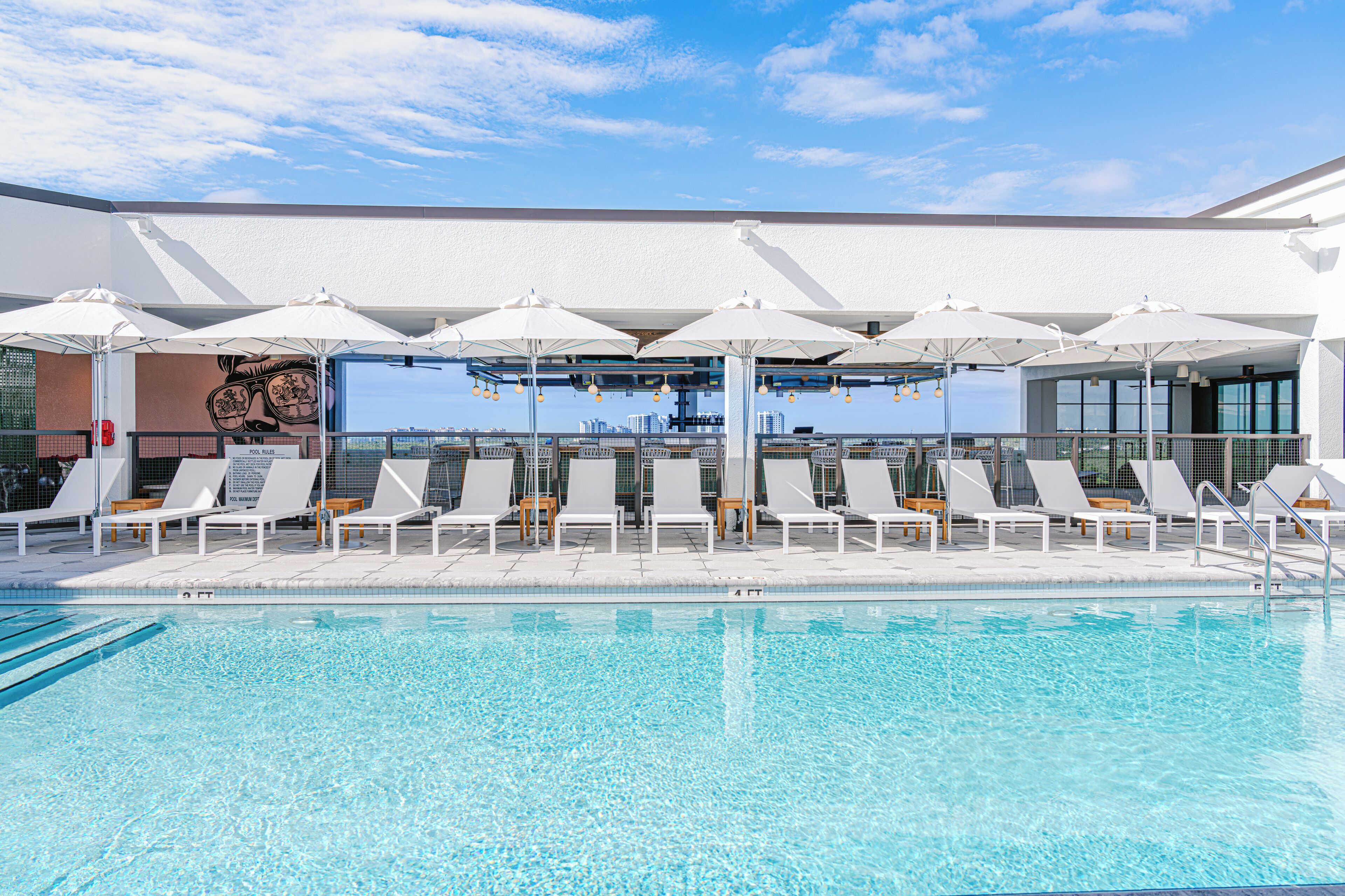 Un área de piscina con sillas de descanso blancas y sombrillas, cielo despejado y vista de edificios al fondo.