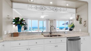 A kitchen with white cabinets, a stainless steel sink, and a dishwasher. There is a large window with a view of the ocean. - Hidden Dunes 402 (Panama City Beach)