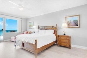 Ein Schlafzimmer mit Bett, Nachttischen, einem Schminktisch und einem großen Fenster mit Blick aufs Meer.
