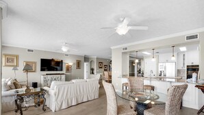 Un salon spacieux avec une table à manger en verre, des chaises en osier, un canapé blanc et un ventilateur de plafond.