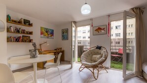 Una sala de estar moderna con comedor, una silla blanca, una estantería y un amplio ventanal con vistas a un edificio y un jardín.