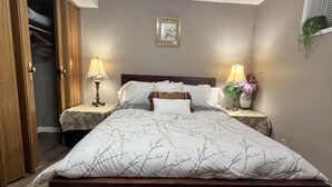 Un dormitorio con una cama grande, un armario de madera, dos mesitas de noche con lámparas, un cuadro en la pared y una ventana pequeña con una planta.