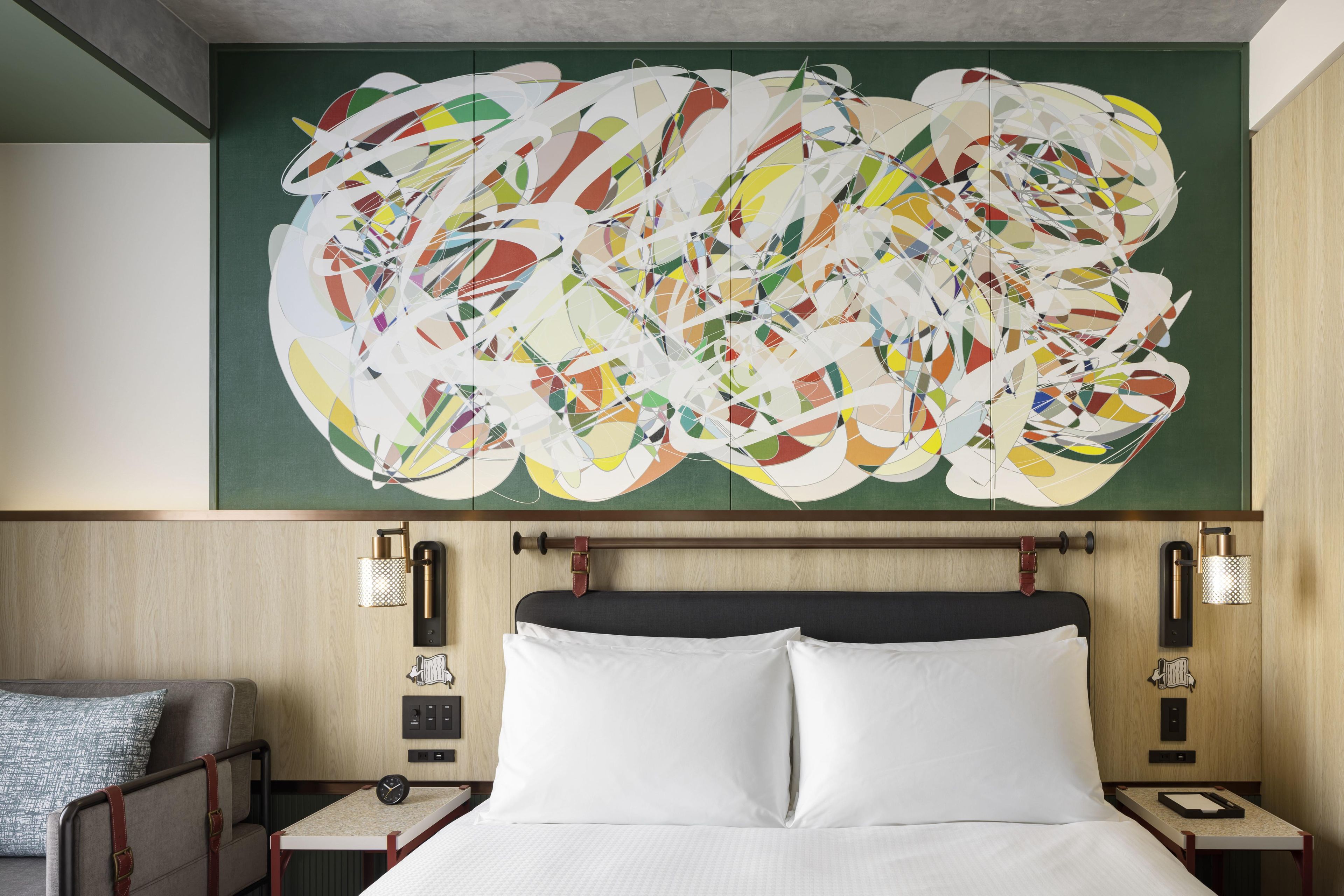 A hotel room with a large abstract painting, a bed with white linens, a wooden headboard, bedside tables with lamps, and a small seating area with a sofa and a patterned throw pillow.