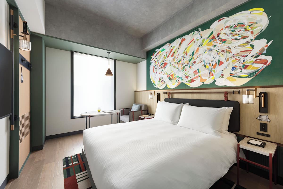 Una habitación de hotel moderna con una cama grande, un escritorio, una silla y un cuadro abstracto de gran tamaño en la pared.