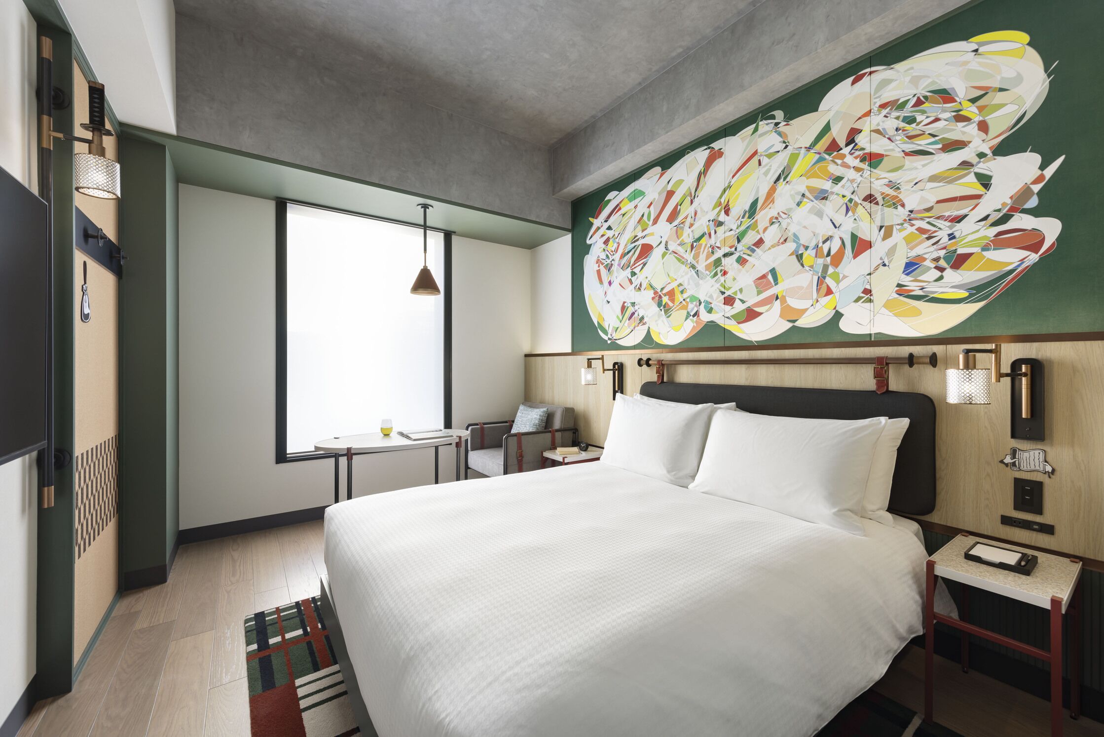 A hotel room with a large bed, a desk, a chair, a window, and a colorful abstract painting on the wall.