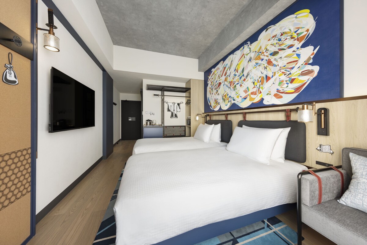 A hotel room with a large bed, a television mounted on the wall, a painting above the bed, and a small kitchenette in the background.