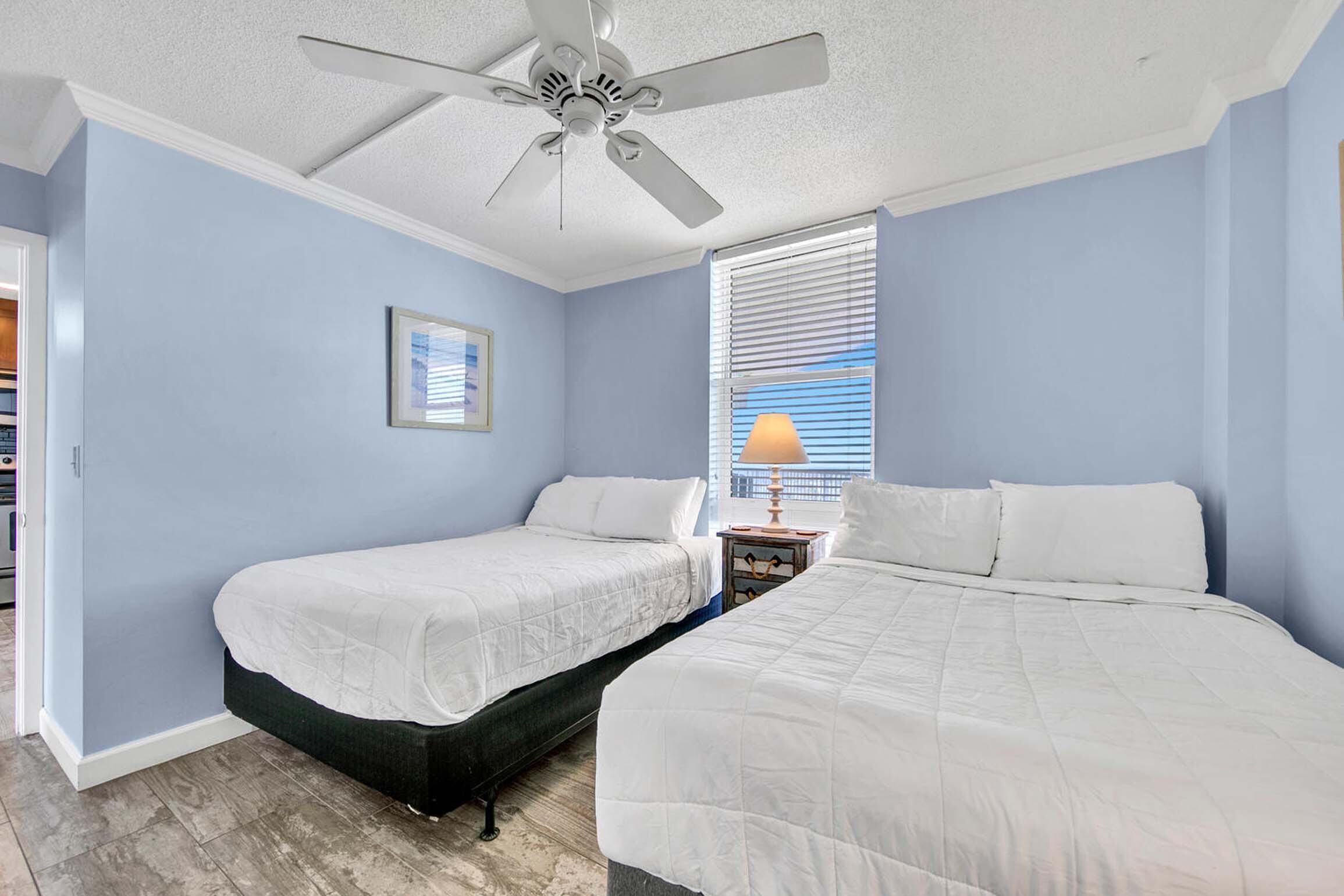 A bedroom with two beds, a ceiling fan, a window with blinds, and a nightstand with a lamp.