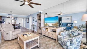 A spacious living room with a large flat-screen TV, a ceiling fan, and a view of the beach through the window. - Dunes of Panama 2002e (Panama City Beach)