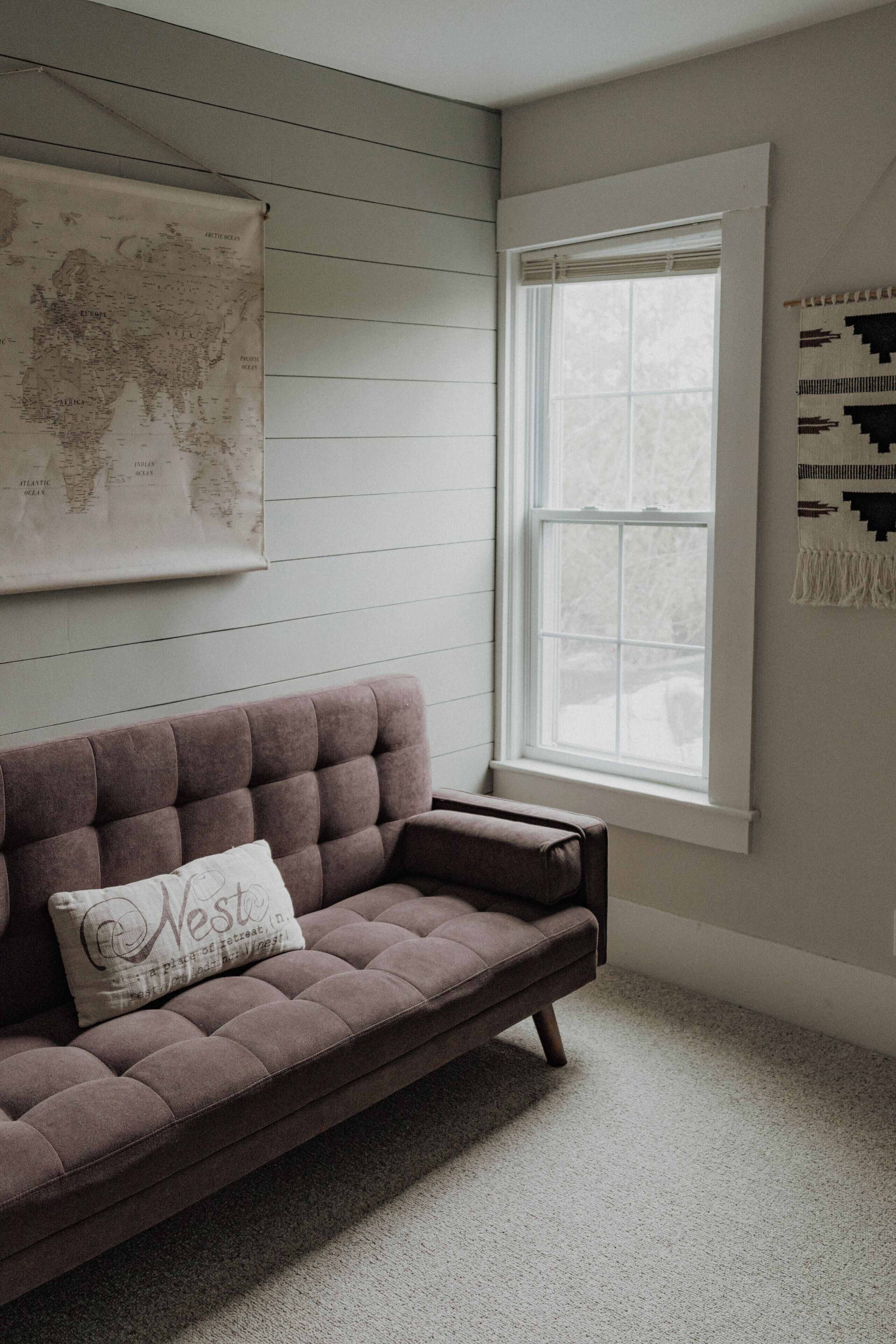 Une pièce avec un canapé marron, une carte du monde accrochée au mur, une fenêtre avec des stores et une décoration murale à motifs.