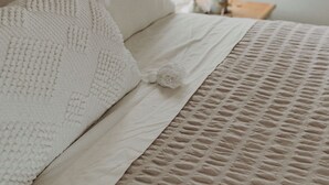 Un lit bien fait, recouvert d’une couverture blanche à texture intéressante et agrémenté d’oreillers, avec une tête de lit en bois, une lampe de chevet et une petite plante posée sur une table de chevet en bois.