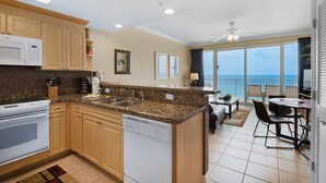 A modern kitchen with granite countertops, wooden cabinets, and a dishwasher. It features a large island with a sink, a built-in oven, and a microwave. The kitchen opens to a balcony with a view of the ocean. - Boardwalk 805 (Panama City Beach)