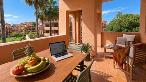 Un balcón con una computadora portátil, una mesa de madera con fruta y mobiliario exterior.