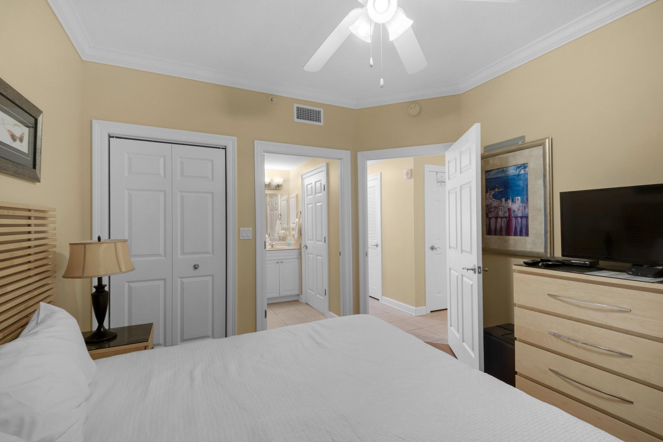 Une chambre à coucher comprenant un lit, une table de chevet avec une lampe, une commode, un téléviseur et un ventilateur de plafond.