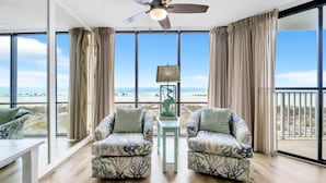 A room with a large window, two patterned armchairs, a small table, and a ceiling fan.