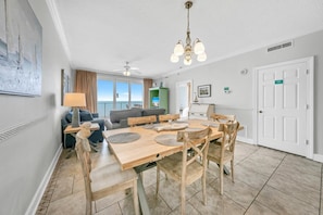 A dining area with a wooden table and chairs, a ceiling fan, and a view of the ocean through large windows. - Twin Palms 804 (Panama City Beach)