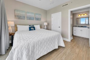 A bedroom with a large bed, a nightstand, a lamp, and a door leading to a bathroom with a vanity and mirror. - Twin Palms 1401 (Panama City Beach)