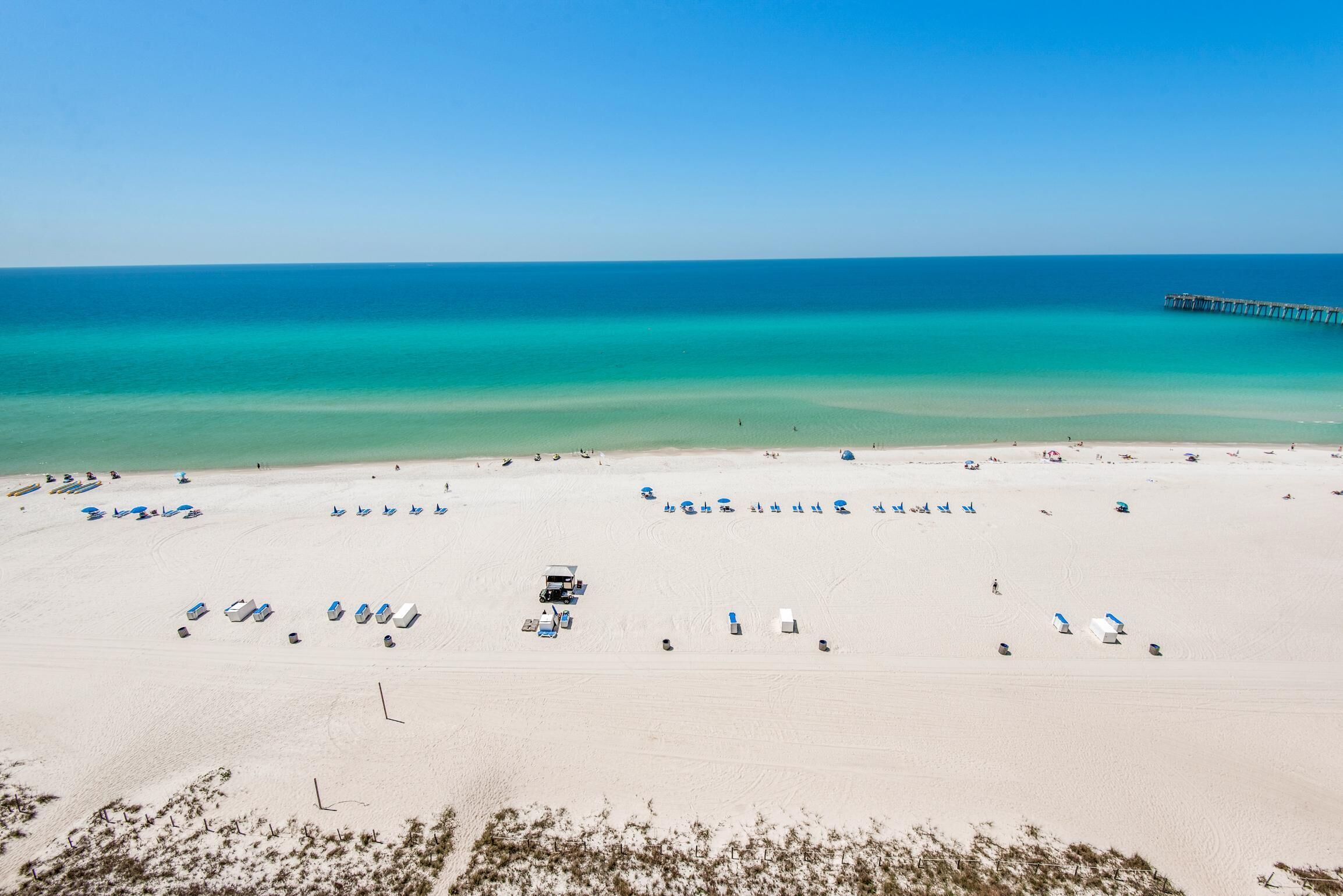 A wide sandy beach with clear turquoise water, a pier extending into the sea, and several beach umbrellas and lounge chairs.