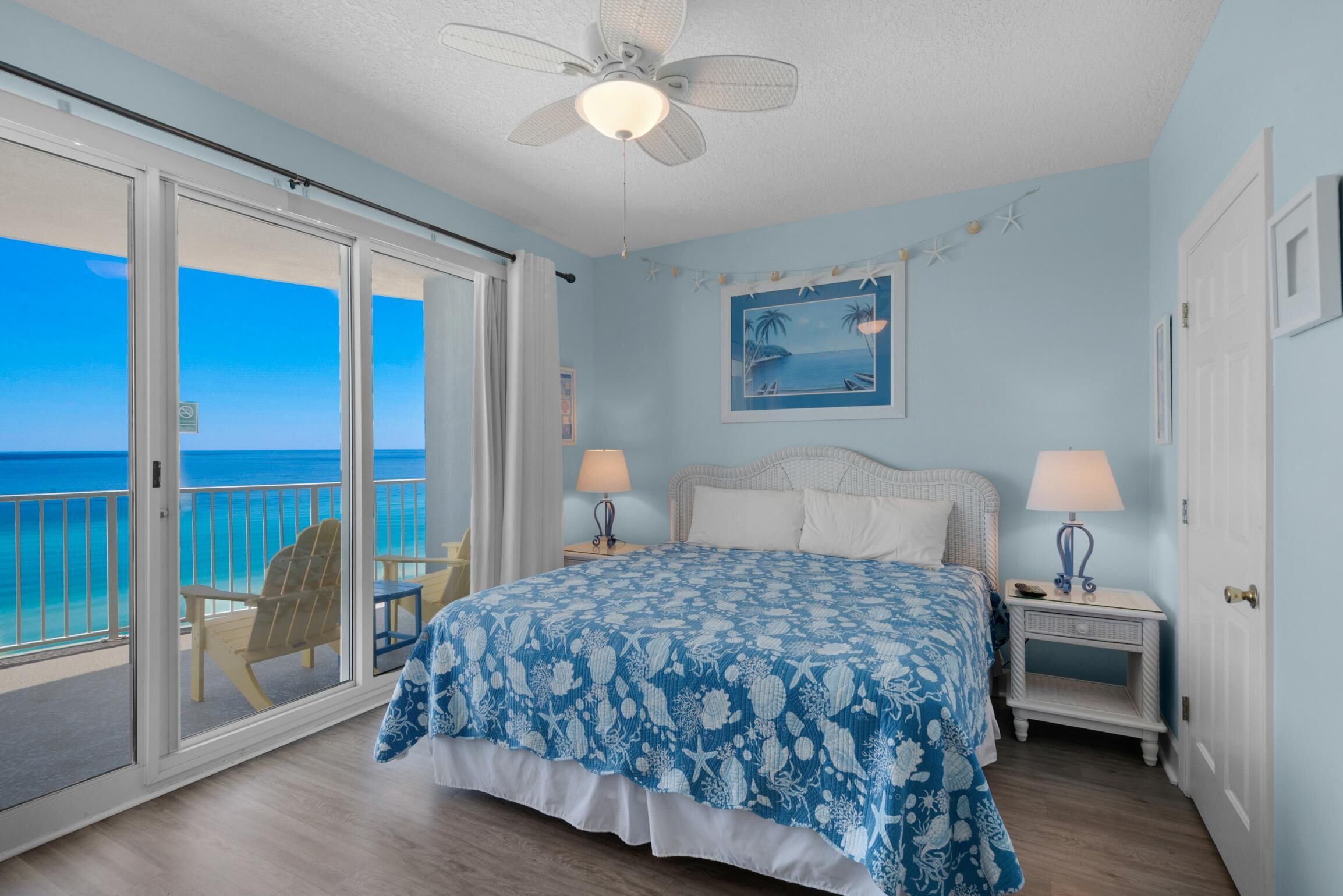 A bedroom with a bed, two bedside tables with lamps, a ceiling fan, a large window with a view of the ocean, and a framed picture on the wall.