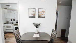 Un coin repas avec une table, des chaises et un vase contenant des plantes séchées. Deux tableaux abstraits encadrés ornent le mur.