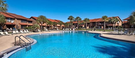 A swimming pool with lounge chairs and palm trees, surrounded by multi-story buildings with red roofs.
