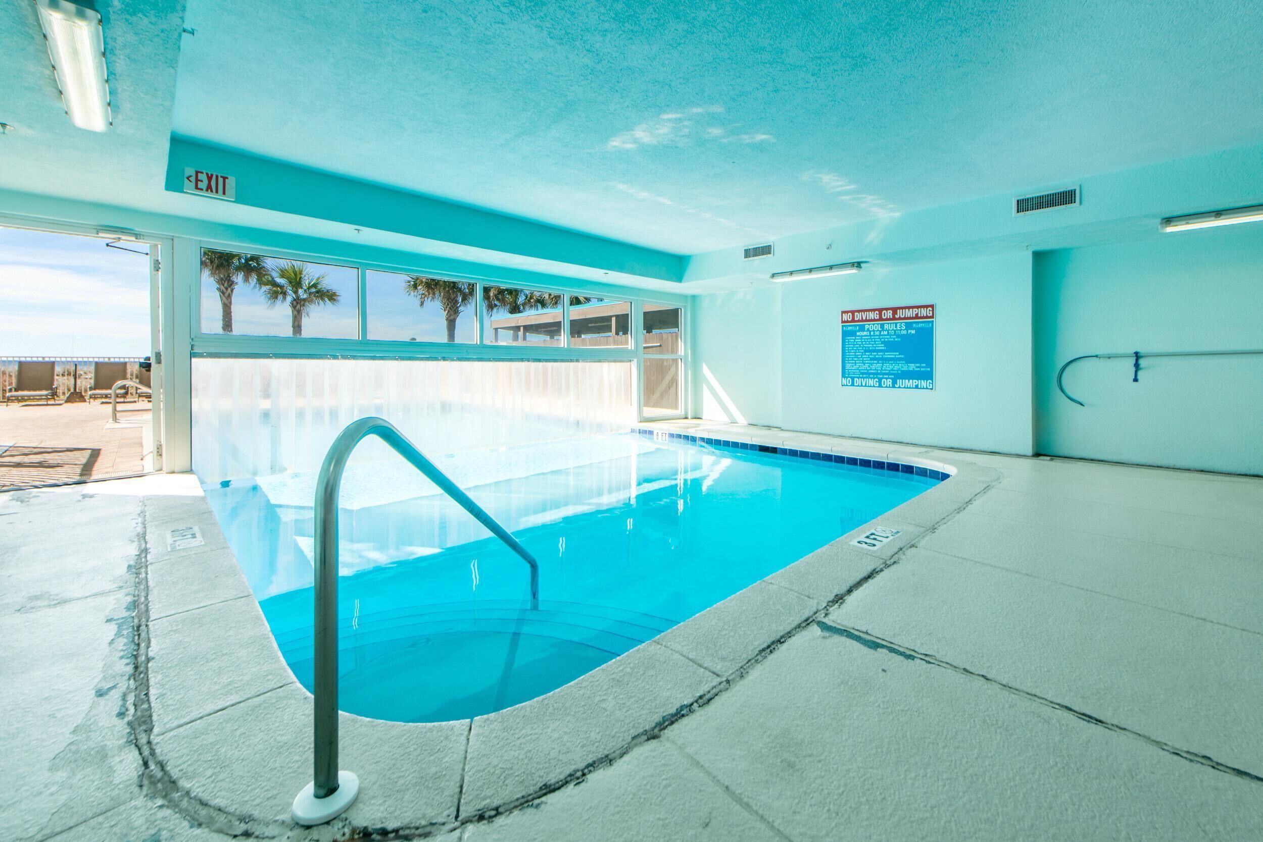 An indoor swimming pool with a clear view of the outside, a sign prohibiting diving or jumping, and a visible exit sign.