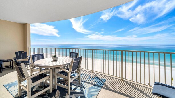 A balcony with a round table and chairs, overlooking a beach with clear blue water and a sandy shore. - Ocean Villa 802 (Panama City Beach)