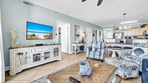 A spacious living room with a large flat-screen TV, a ceiling fan, and a kitchen area with wooden cabinets and a dining table. - Ocean Villa 802 (Panama City Beach)