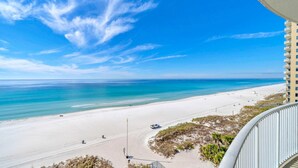 A beach view from a balcony with a clear sky, a sandy beach, and a building in the background. - Ocean Villa 802 (Panama City Beach)