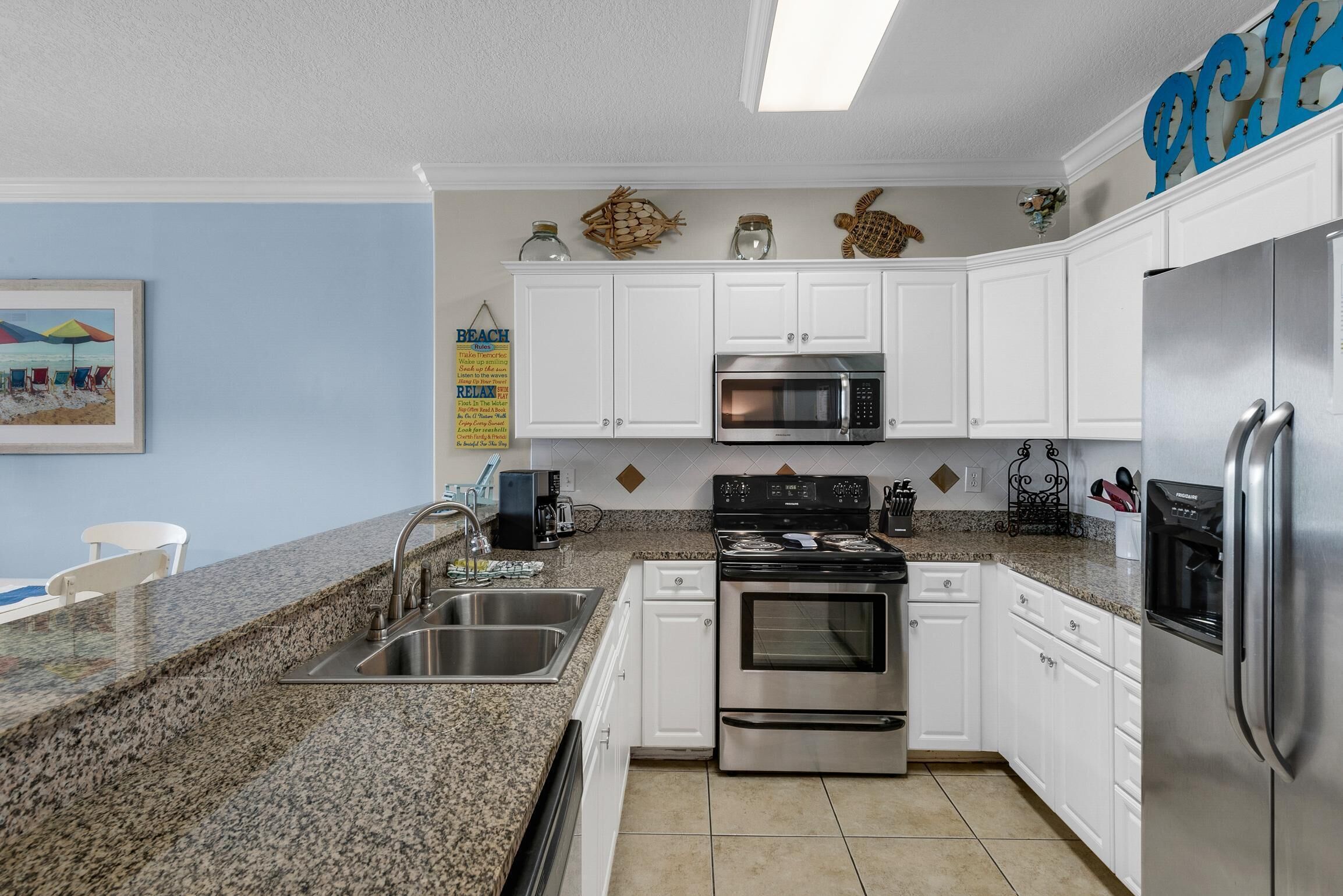 Cocina con armarios blancos, encimera de granito, electrodomésticos de acero inoxidable y detalles decorativos como una tortuga y una escena de playa.
