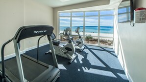 Ein Fitnessraum mit Laufband, Crosstrainer und Ergometer, mit Blick auf einen Strand.