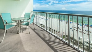 Ein Balkon mit zwei grünen Stühlen und einem kleinen Tisch, der einen Blick auf einen Strand mit Sonnenschirmen und Menschen bietet.