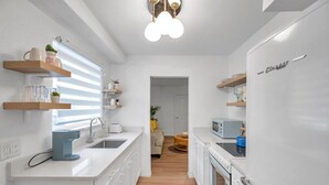 Et moderne køkken med hvide skabe, en indbygget ovn og en mikrobølgeovn. Der er træhylder over disken og en loftlampe.