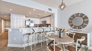 A modern kitchen with white cabinets, a central island with stools, and a dining area with a round table and chairs. - Majestic Beach Towers 2-2003 (Panama City Beach)