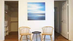 Een gang met twee houten stoelen en een kleine ronde tafel, een schilderij aan de muur en een witte deur.