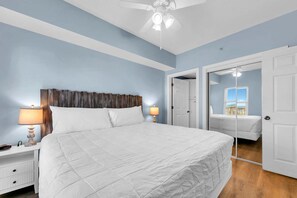 Une chambre avec un grand lit, une table de chevet avec une lampe, un ventilateur de plafond et une porte donnant sur une autre pièce.