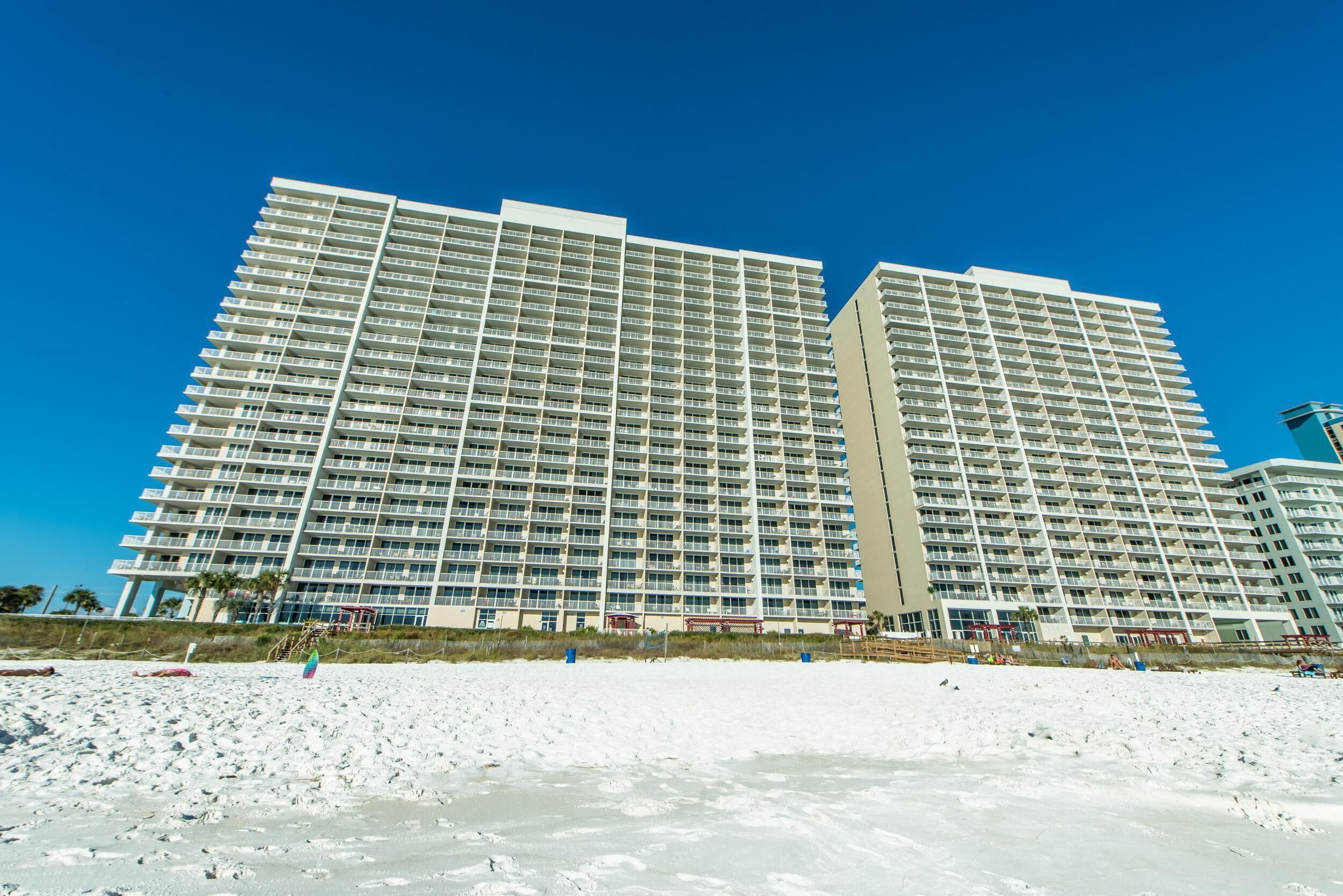 Ein Hochhaus-Hotel mit mehreren Balkonen, ein Strand im Vordergrund und strahlend blauer Himmel.