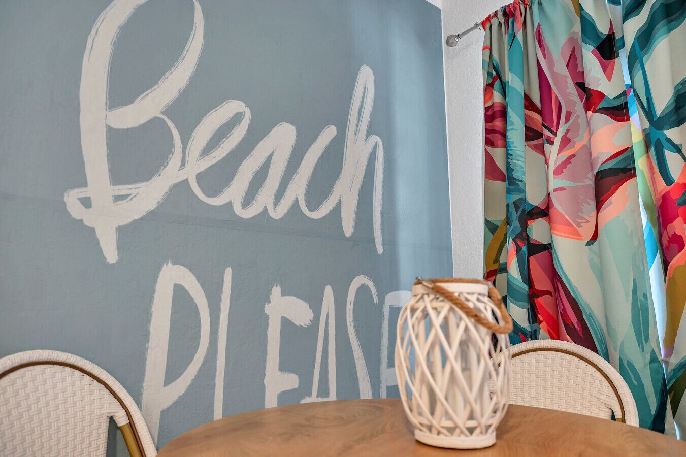 A room with a wall painted in a light blue color and the words "Beach" and "Please" written on it. There is a wicker chair and a wooden table in the foreground.