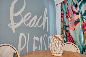 Ein Zimmer, dessen Wand in Hellblau gestrichen ist und die Wörter "Beach" und "Please" zeigt. Im Vordergrund steht ein Korbstuhl und ein Holztisch.