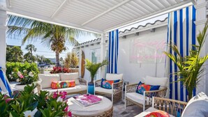 An outdoor seating area with wicker furniture, colorful cushions, and a neon sign on the wall.