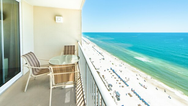 A balcony with a glass table and chairs, overlooking a beach with people and umbrellas. - Majestic Beach Towers 1-1706 (Panama City Beach)