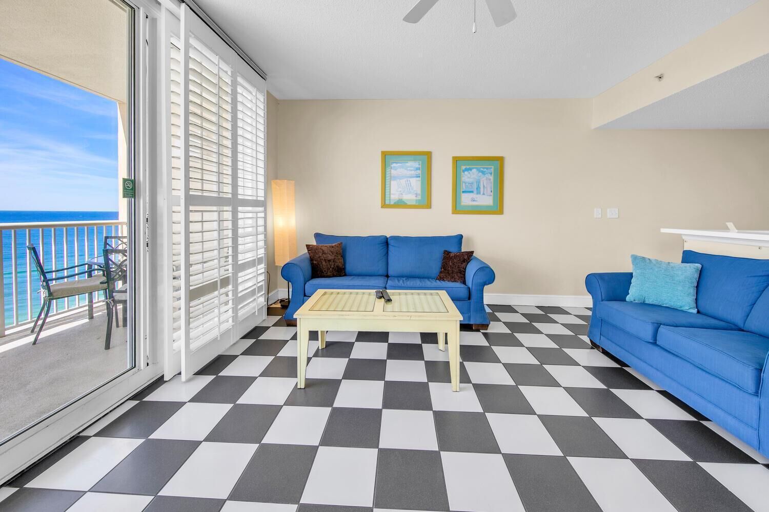 Una habitación con piso a cuadros, dos sofás azules, una mesita y un balcón con vista al océano.