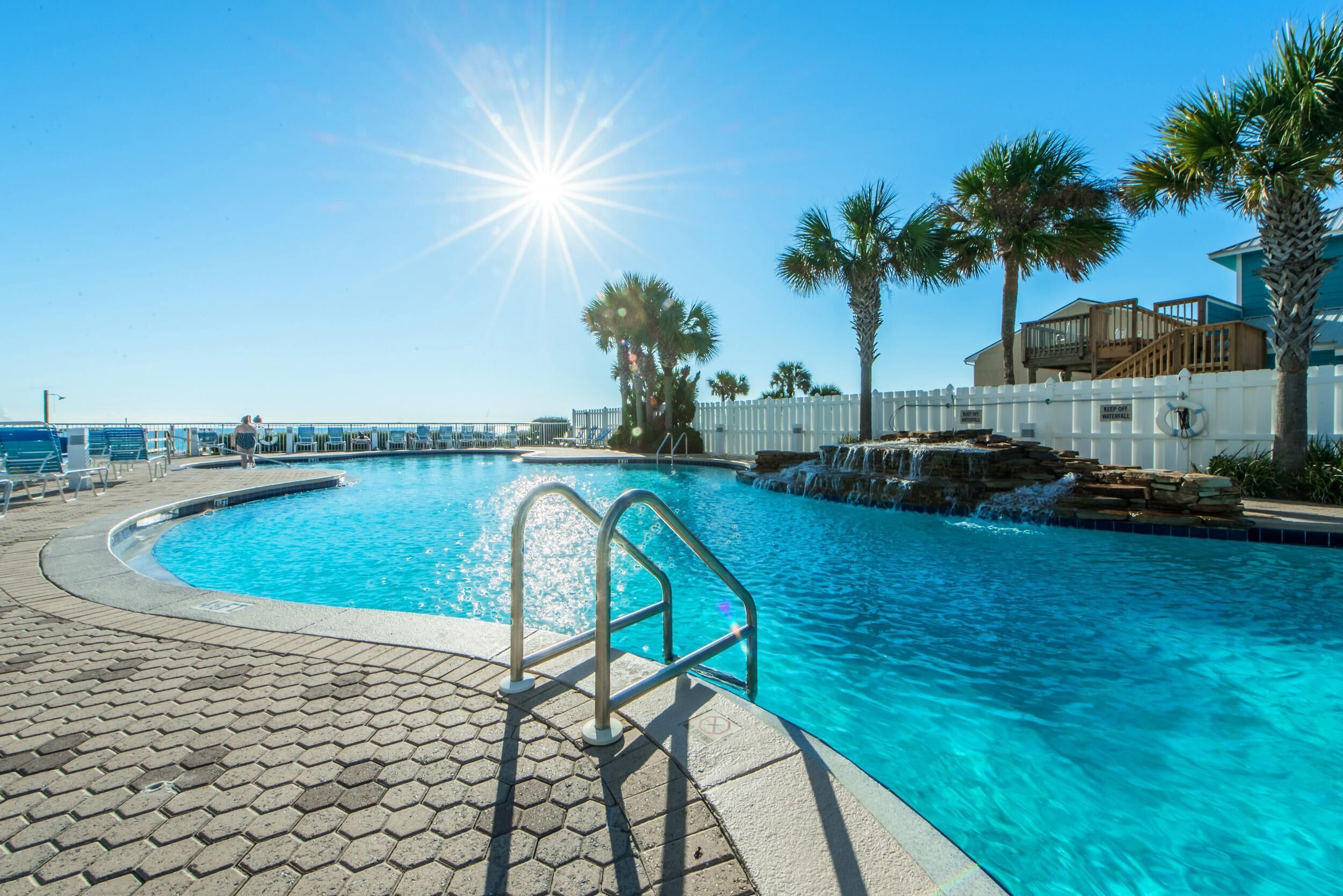 A swimming pool with a sunlit sky, palm trees, and a white fence.