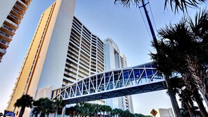 A modern hotel with a prominent glass bridge structure, high-rise buildings in the background, and palm trees lining the street. - Majestic Beach Towers 1-1006 (Panama City Beach)