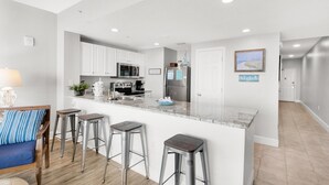 A modern kitchen with white cabinets, stainless steel appliances, and a marble island with bar stools. - Majestic Beach Towers 1-1006 (Panama City Beach)
