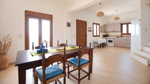 A spacious dining area with a wooden table and chairs, a kitchen in the background, and a staircase leading to an upper floor. - Angelika House -Amazing Sea view (Faistos)