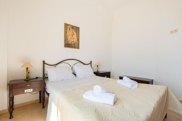 A neatly made bed with white linens and two rolled towels, a nightstand with a lamp, and a painting on the wall. - Angelika House -Amazing Sea view (Faistos)
